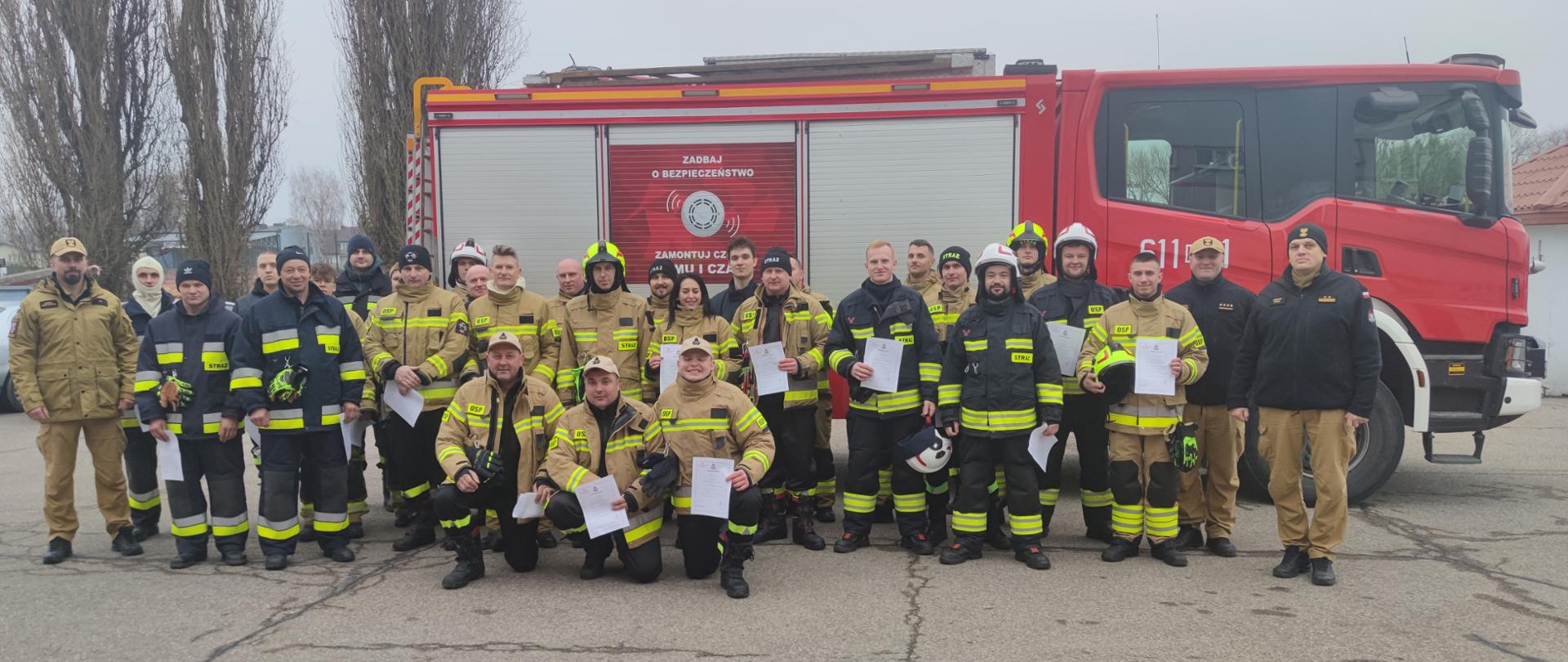 Zdjęcie grupowe przedstawia oddział strażaków pozujących do oficjalnego zdjęcia na placu przed czerwonym wozem strażackim.
Grupa liczy około dwudziestu pięciu osób, w większości w pełnym umundurowaniu bojowym w kolorze piaskowym/żółtym i czarnym. Kilku strażaków klęczy z przodu, trzymając w rękach białe dokumenty, co sugeruje uroczystość ukończenia szkolenia lub wręczenie certyfikatów.