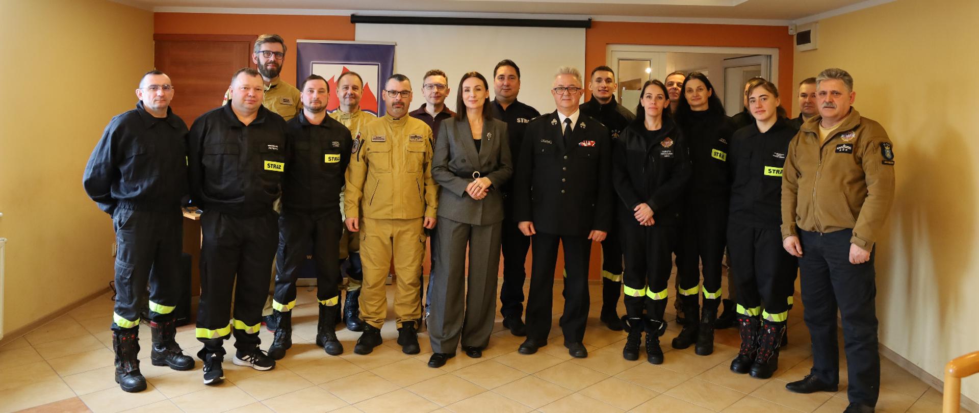 grupa osób, w tym wojewoda i strażacy, stoją w sali i pozują do zdjęcia