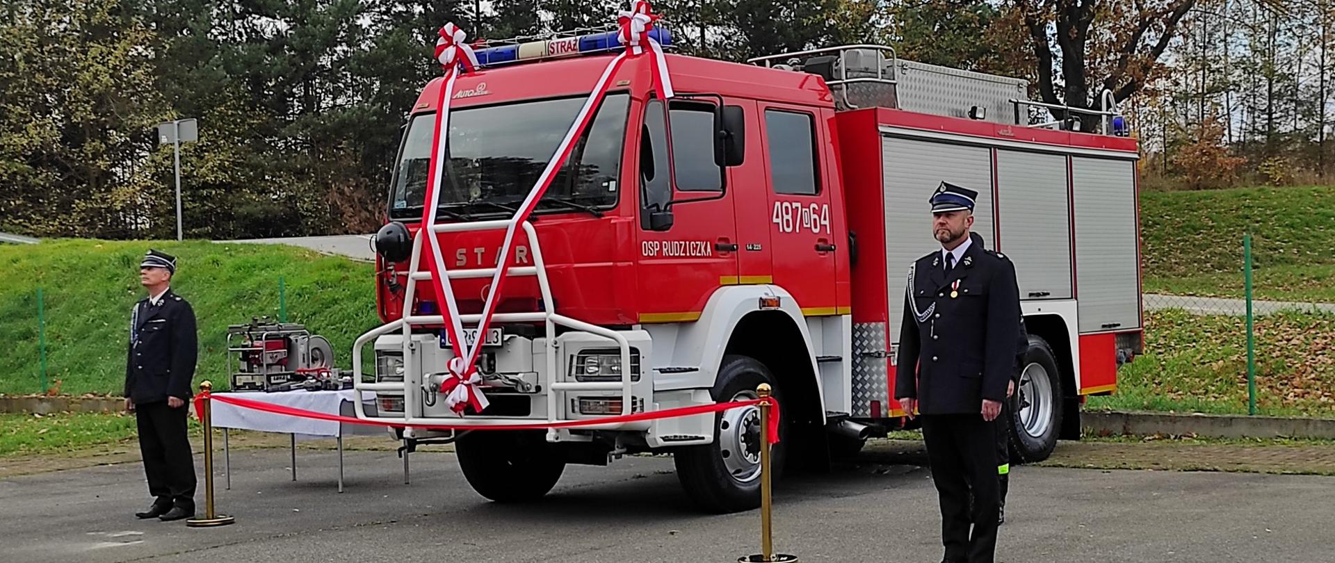 Zdjęcie przedstawia udekorowany samochód ratowniczo gaśniczy straży pożarnej oraz sprzęt hydrauliczny ustawiony na stoliku. Przed samochodem wstęga koloru czerwonego. Po bokach stoją strażacy w mundurach galowych