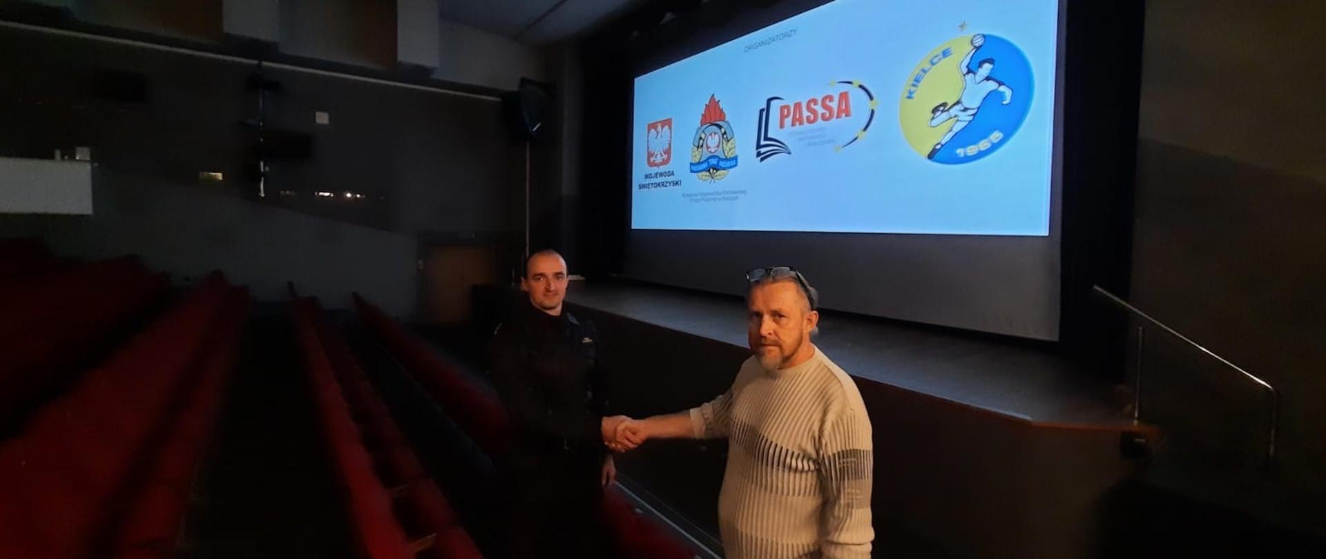 Dwóch mężczyzn w kinie podających sobie prawe dłonie. Jeden z nich (strażak) w umundurowaniu dowódczo-sztabowym. Na ekranie wyświetlany spot informacyjny dotyczący zagrożeń związanych z sezonem grzewczym.