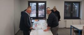 Na zdjęciu widać grupę dorosłych osób stojących wokół stołu, na którym rozłożone są kolorowe prace plastyczne. Uczestnicy pochylają się nad rysunkami, uważnie je oglądając i omawiając ich treść.
