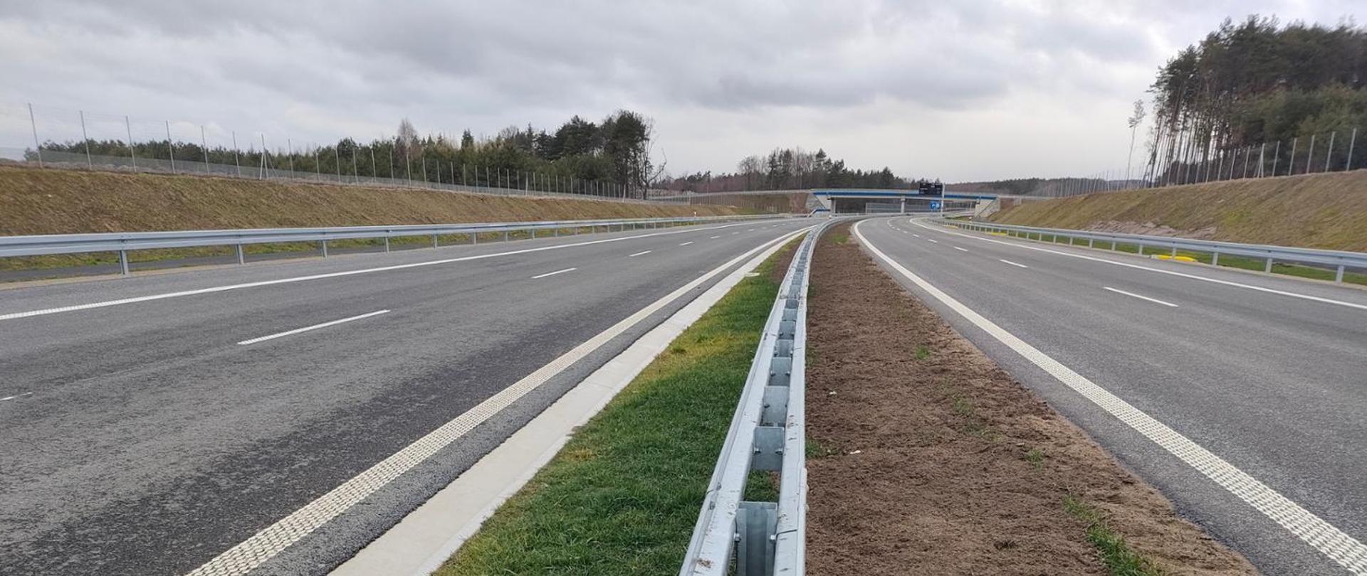 Widok na drogę dwupasmową rozdzieloną pasem zieleni i barierami energochłonnymi. W tle widać wiadukt nad drogą.