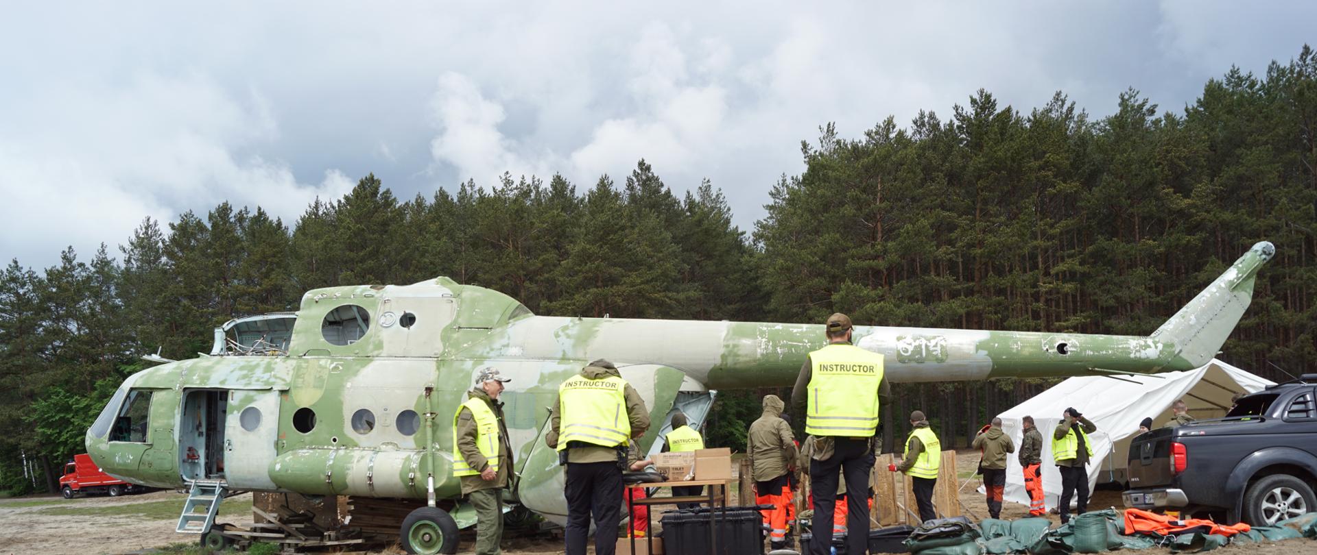 na zdjęciu pokazani są studenci ratownictwa medycznego i adepci zawodu ratownika medycznego oraz ich instruktorzy, którzy na tle śmigłowca i namiotowego stanowiska operacyjnego szykują się do dalszych ćwiczeń