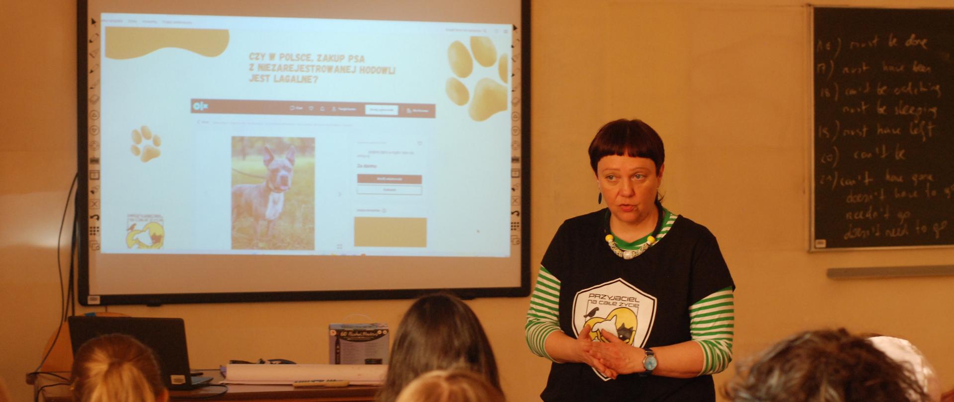Kobieta prowadzi prelekcję w sali lekcyjnej przed grupą słuchaczy siedzących w ławkach. Za nią ekran z prezentacją dotyczącą legalności zakupu psa w Polsce