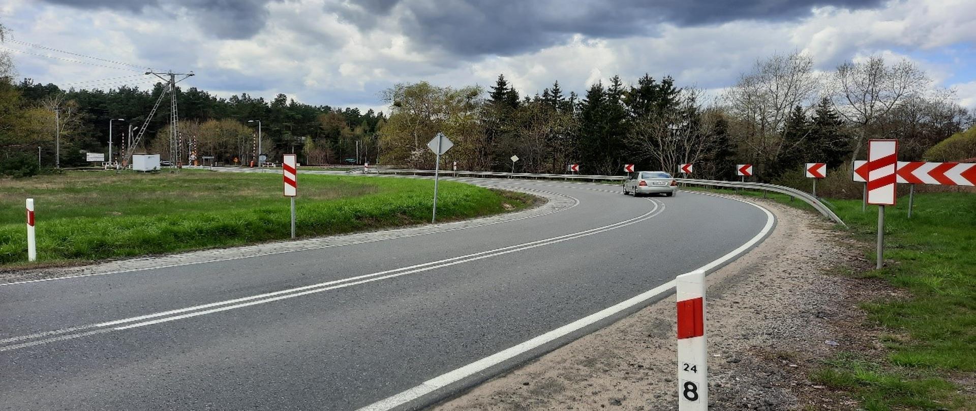 Odcinek drogi o charakterystycznym ostrym zakręcie w prawo, zabezpieczonym barierą energochłonną oraz oznakowaniem ostrzegawczym w postaci znaków kierunkowych (czerwone strzałki na białym tle). Na pierwszym planie znajduje się biały słupek drogowy z oznaczeniem kilometrażu (24/8). Całość otacza teren zielony.