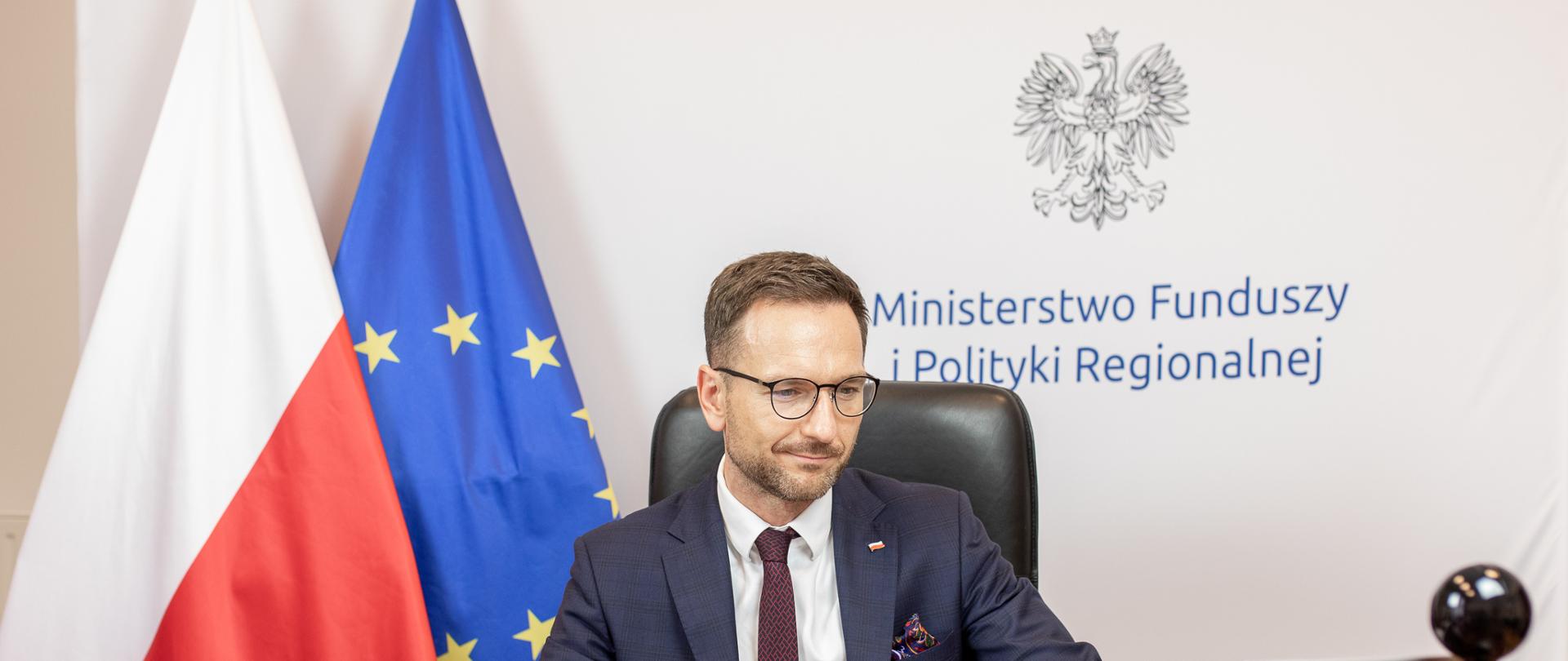 Minister Waldemar Buda przy biurku, w tle flaga Polski i Unii Europejskiej