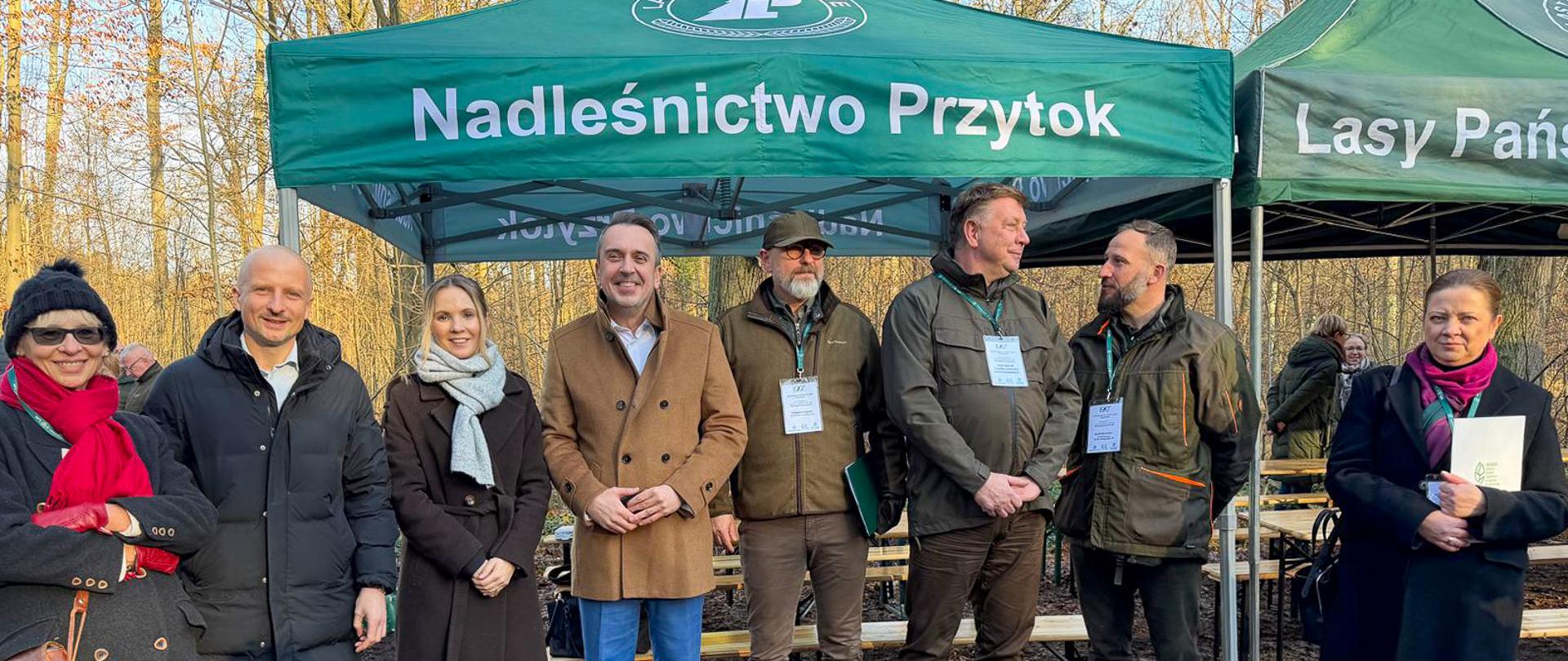 Na zdjęciu widać grupę ośmiu dorosłych osób stojących na zalesionym, pokrytym ziemią i liśćmi terenie. Za osobami znajdują się zielone namioty, widnieje na nich napis: "Nadleśnictwo Przytok", "Lasy Państwowe" Osoby stoją w rzędzie, z których większość jest ubrana w ciepłe, zimowe okrycia.