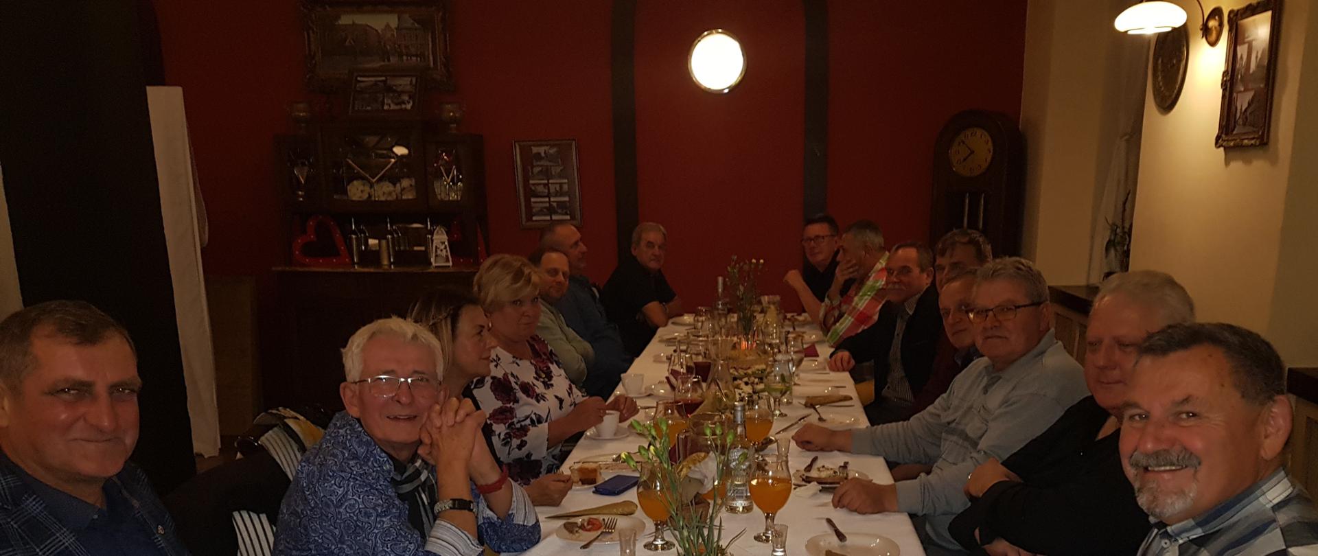 Zdjęcie przedstawia stół wokół którego siedzą mężczyźni. Stół zastawiony jest jedzeniem oraz napojami.