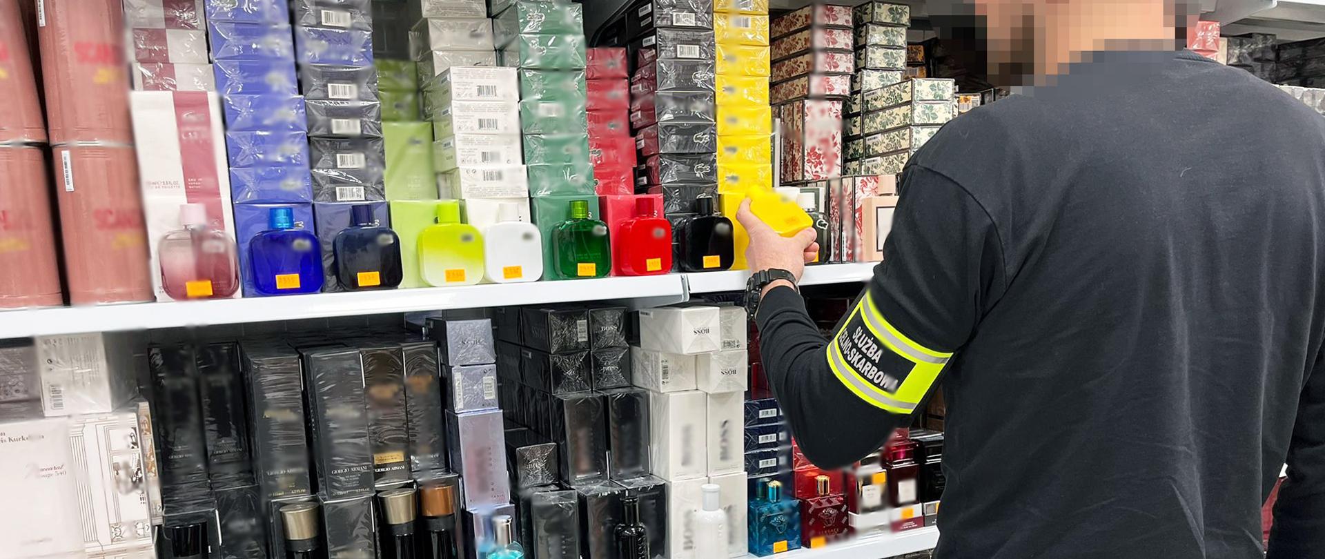 Osoba w odzieży służbowej stoi przed półką z perfumami i trzyma jedno z opakowań.