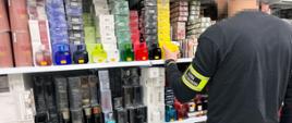 Osoba w odzieży służbowej stoi przed półką z perfumami i trzyma jedno z opakowań.