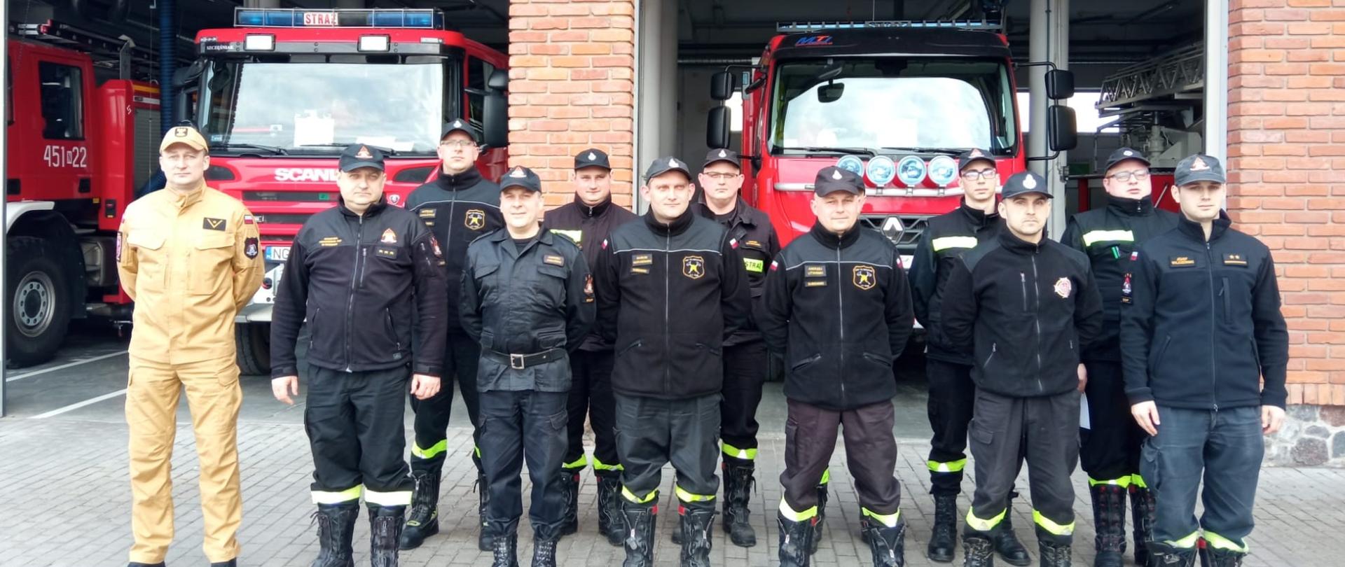 Zdjęcie przedstawia zespół egzaminacyjny oraz uczestników szkolenia kierującego działaniem ratowniczym dla strażaka ratownika Ochotniczych Straży Pożarnych (dowódcy OSP), ustytuowanych na tle pojazdów pożarniczych zlokalizowanych w garażu Komendy Powiatowej Państwowej Straży Pożarnej w Gołdapi