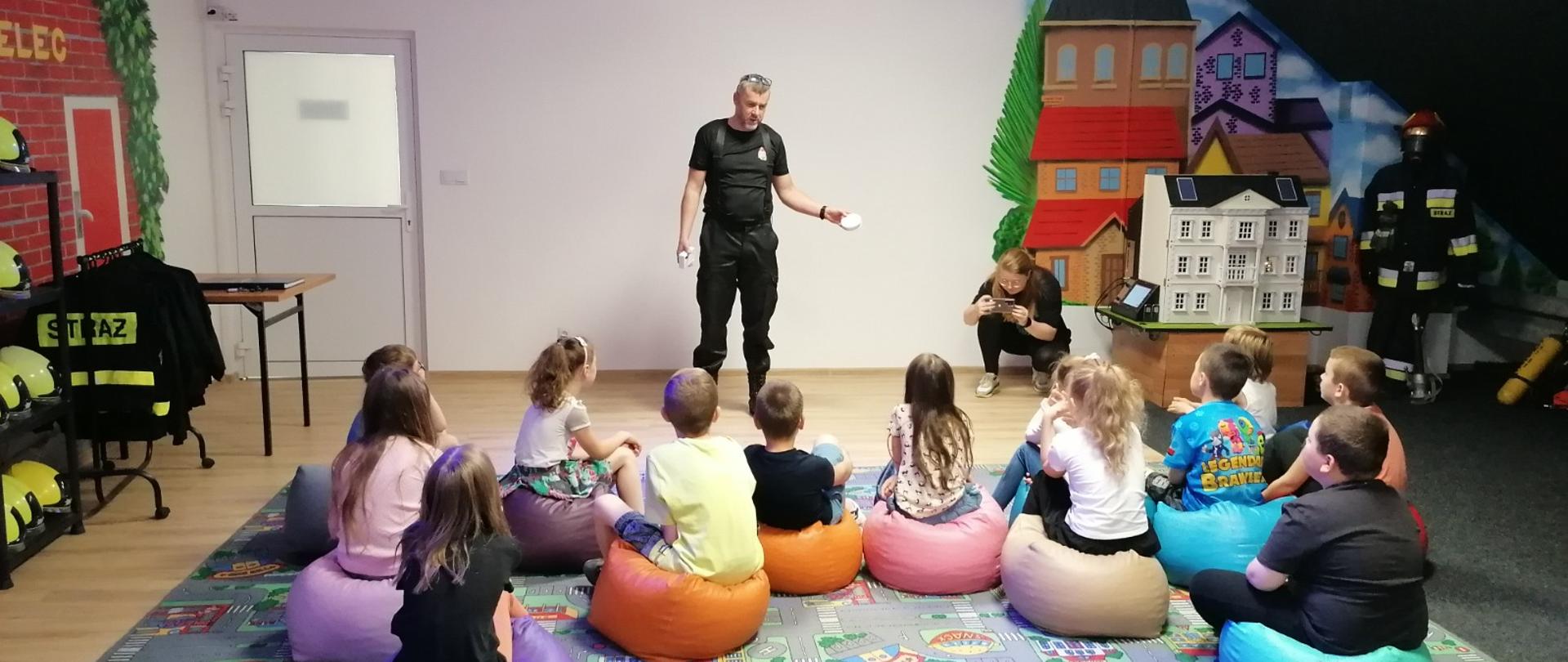 Wnętrze pomieszczenia. Na podłodze położony dywan w kolorowe wzory. Na dywanie znajdują się kolorowe pufy-siedziska, na których siedzi grupka 12 dzieci. Przed dziećmi stoi funkcjonariusz PSP ubrany w czarne spodnie oraz czarną koszulkę. Trzyma w dłoni biały przedmiot - prawdopodobnie czujkę dymu. Funkcjonariusz stoi na tle białej ściany. Z lewej strony w ścianie znajdują się drzwi z częściowym [przeszkleniem. Z lewej strony widać stojak z zabawkowym hełmami w kolorze żółtym oraz wieszak z czarnymi bluzami z napisem STRAŻ na plecach. Z prawej strony widać ubranie specjalne w kolorze granatowym oraz hełm w kolorze czerwonym umieszczone na manekinie oraz zabawkowy domek o 3 kondygnacjach. Przy domku kuca kobieta robiąc zdjęcia dzieci za pomocą telefonu