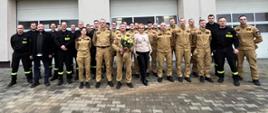 grupa strażaków stojących przed budynkiem strażnicy, jeden strażak z kwiatami pozujący do zdjęcia
