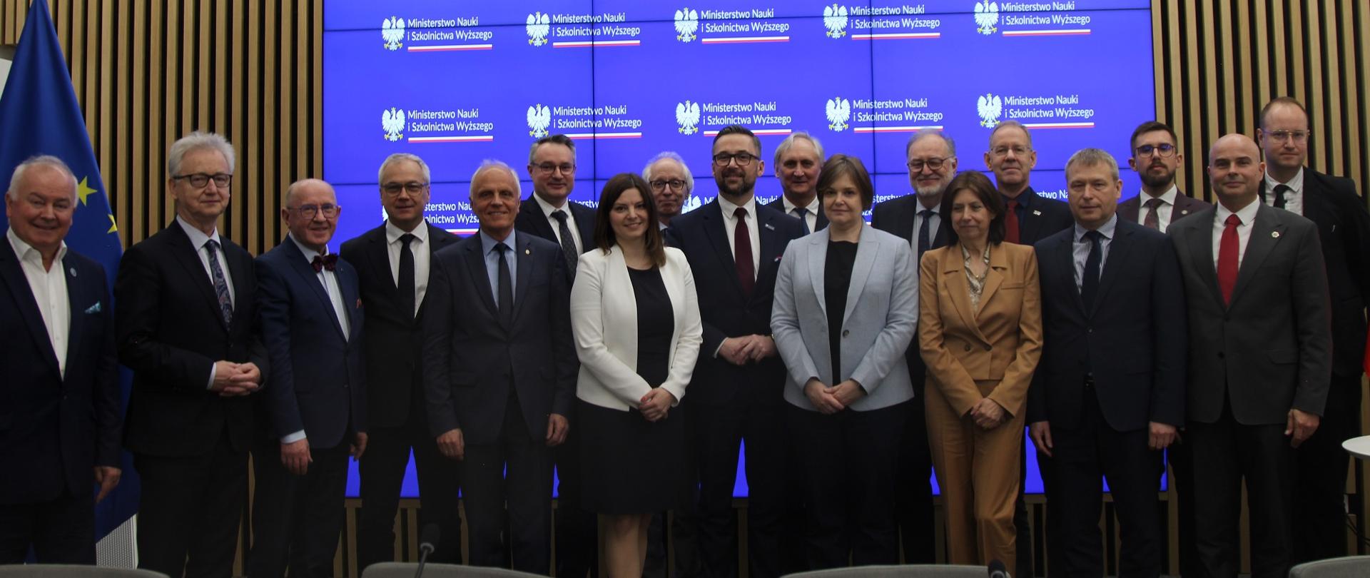 Zdjęcie zbiorowe uczestników spotkania, pod wbudowanym w ścianę z pionowych drewnianych listewek wielkim ekranem z napisem Ministerstwo Nauki i Szkolnictwa Wyższego stoi grupa ok. dwudziestu osób.