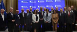 Zdjęcie zbiorowe uczestników spotkania, pod wbudowanym w ścianę z pionowych drewnianych listewek wielkim ekranem z napisem Ministerstwo Nauki i Szkolnictwa Wyższego stoi grupa ok. dwudziestu osób.