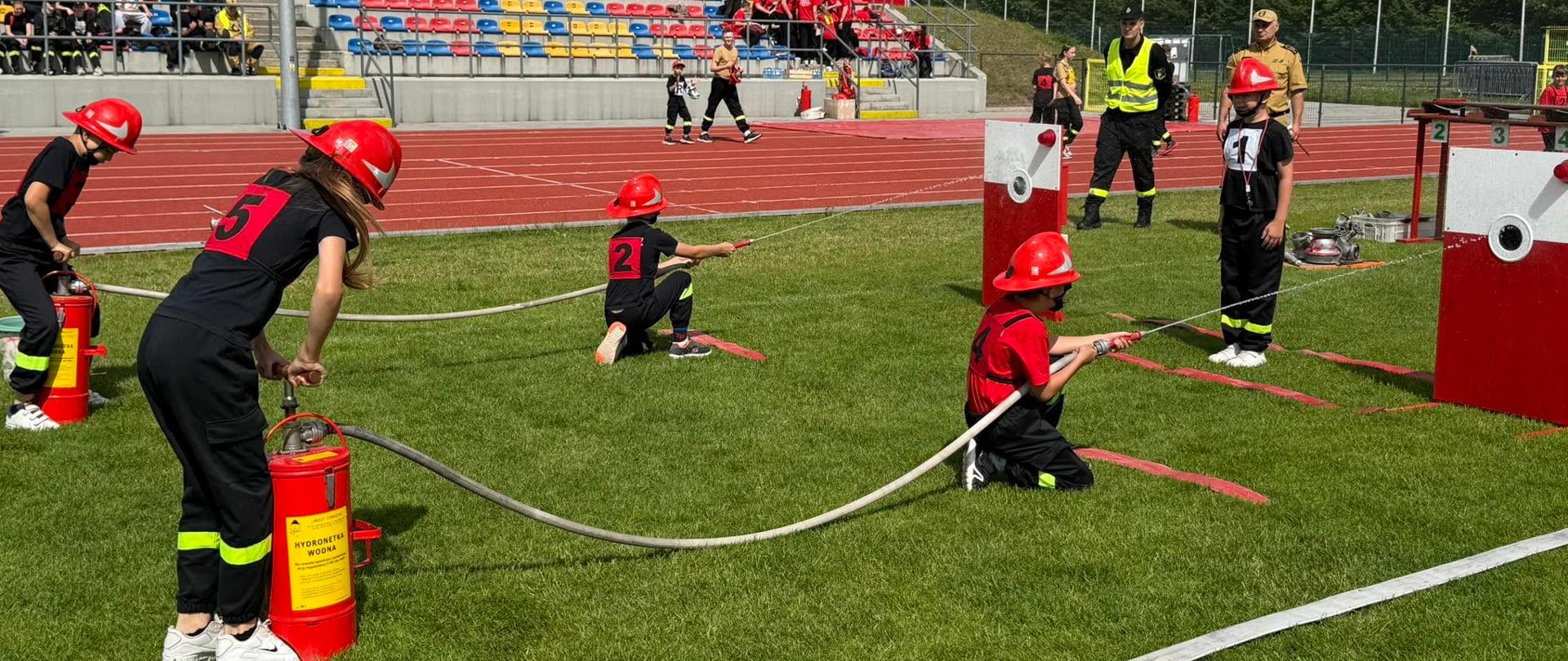 Na zielonej murawie stadionu kilkoro dzieci w czerwonych kaskach i czarnych strojach strażackich wykonuje zadanie z użyciem węży strażackich. Dwoje dzieci klęczy na trawie, kierując strumień wody lub przygotowując sprzęt do celu znajdującego się przed nimi. Inne dzieci stoją obok, obsługując elementy instalacji. Po lewej stronie stoją czerwone pojemniki przypominające gaśnice lub zbiorniki treningowe. W tle znajdują się kolorowe trybuny, obserwatorzy oraz osoby nadzorujące konkurencję. Zdjęcie przedstawia zorganizowane dziecięce zawody pożarnicze na stadionie sportowym.