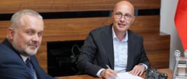 Wojciech Kamiński, Prezes Zarządu PKW S.A. oraz Robert Kropiwnicki, Sekretarz Stanu w MAP siedzą przy stole. Wiceminister podpisuje dokumenty. 