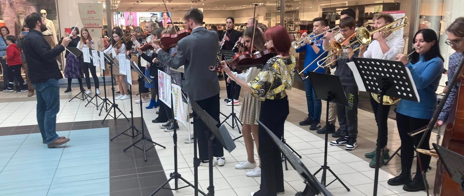 Jakub Unolt dyryguje orkiestrą szkolną, która gra na stojąco, przed sklepem w CH Ferio.