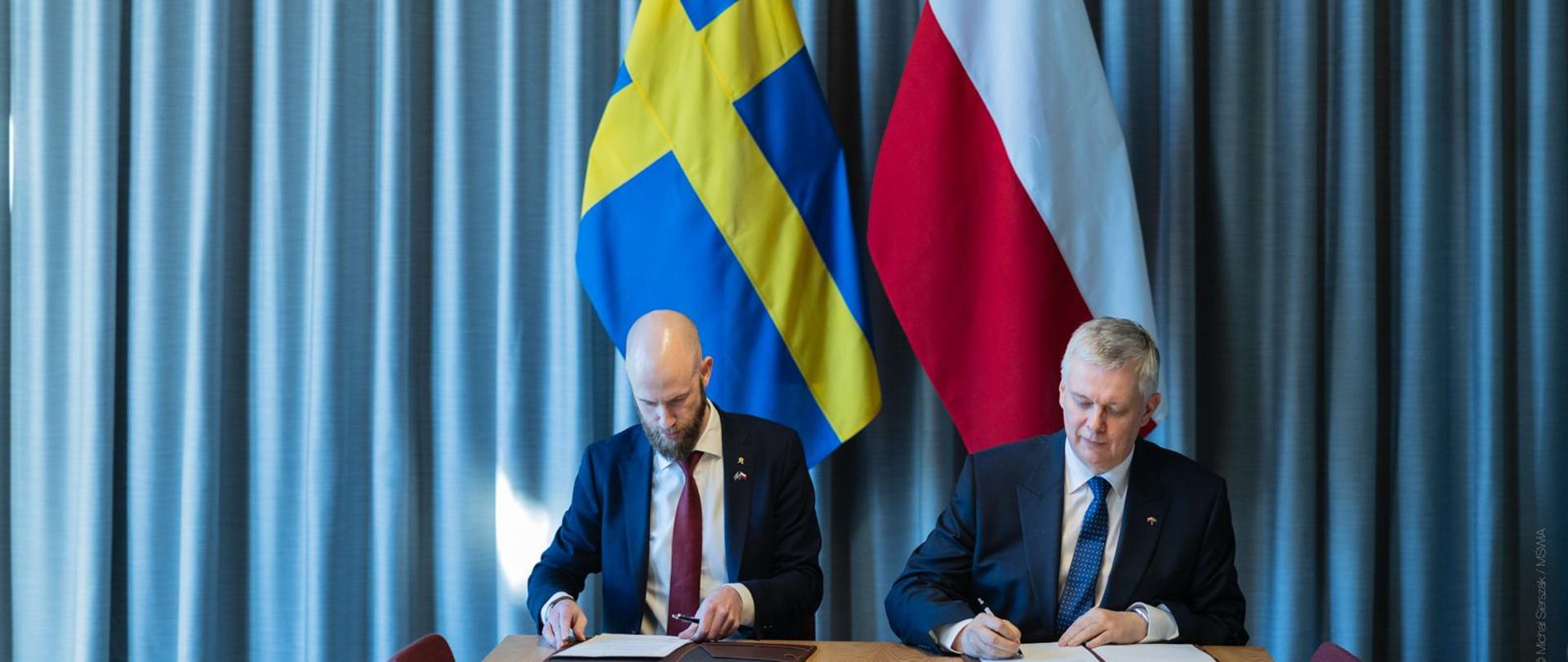 Ministrowie Siemoniak i Bohlin podpisują dokumenty na tle flag polskiej i szwedzkiej 
