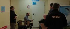 Zdjęcie przedstawia funkcjonariuszy z Komendy Powiatowej PSP w Tucholi (w umundurowaniu koszarowym oraz w ubraniu cywilnym), czekających w kolejce do punktu szczepień przeciwko COVID-19.
