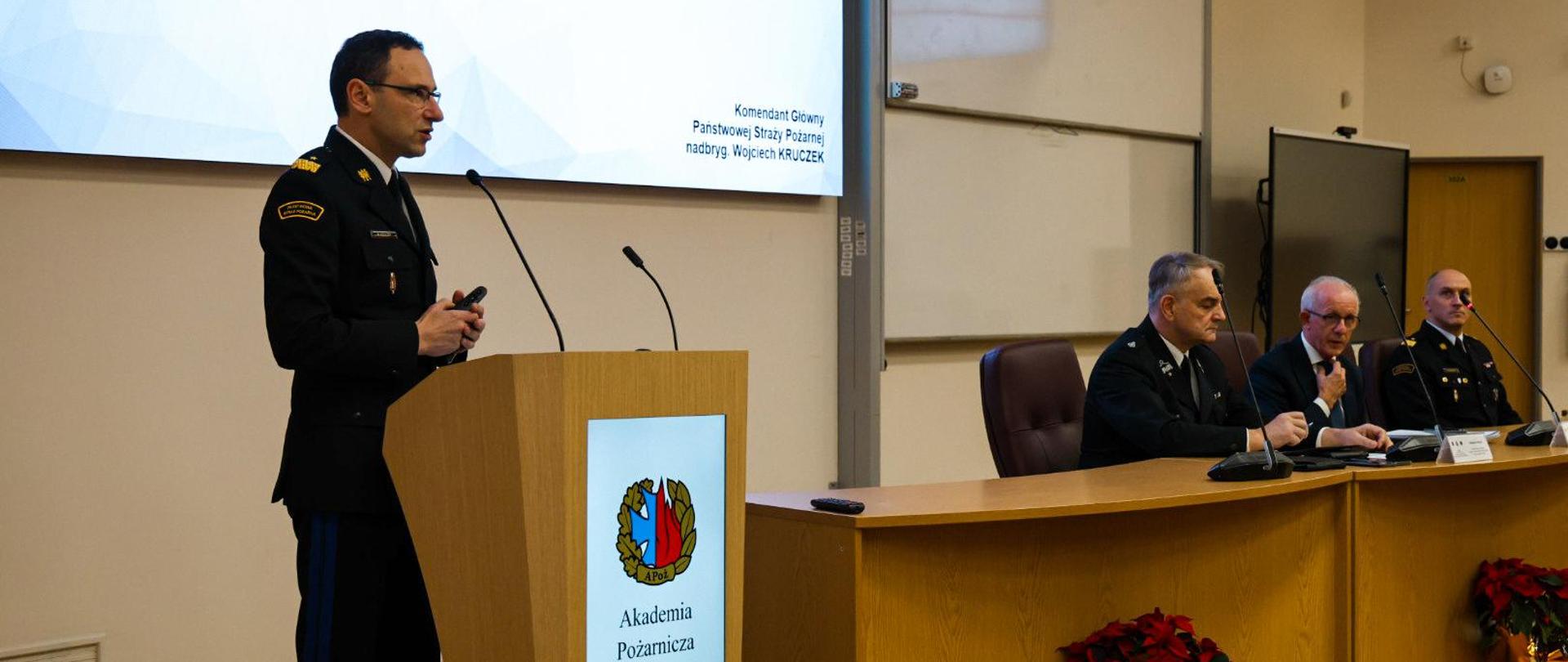 Oficer PSP w mundurze galowym wygłasza prezentację na temat współpracy PSP i OSP; na sali siedzą inni mundurowi oraz cywil, a na ekranie widać slajd z tytułem wystąpienia.