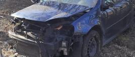 Zdjęcie przedstawia zniszczony samochód osobowy znajdujący się w pasie drogi wojewódzkiej, który brał udział w zdarzeniu. Pojazd posiada rozbity przód oraz uszkodzoną przednią szybę. 