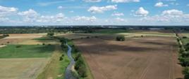 Zdjęcie lotnicze przedstawiające wąską rzekę płynącą pomiędzy rozległymi polami uprawnymi. W oddali widoczna jest zabudowa miejska, a nad krajobrazem znajdują się białe chmury