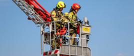 Na zdjęciu widoczni w koszu podnośnika hydraulicznego na wysokości podczas rozpoznania strażacy jeden z Państwowej Straży Pożarnej i drugi z Ochotniczych Straży Pożarnych podczas ćwiczeń taktyczno-bojowych na zakładzie TLC Ocynkownia & Packing Center w Gorlicach. Strażacy ubrani w ubrania bojowe koloru piaskowego w hełmach jeden w czerwonym drugi w białym na tle nieba. 