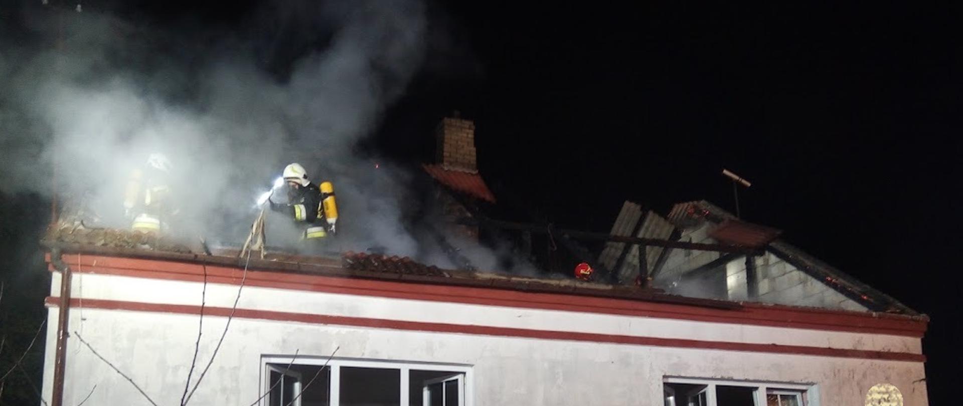 Budynek mieszkalny po pożarze. Pokrycie dachu uległo zniszczeniu. Nad budynkiem widoczny dym. Na dachu pracujący strażak. Wyposażony jest w aparat ochrony układu oddechowego.