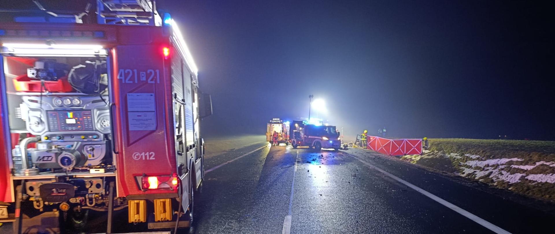 Jest pora nocna. Na zdjęciu widać samochody pożarnicze oświetlające teren działań. W tle widać karetkę pogotowia oraz parawan zasłaniające miejsce udzielania pomocy medycznej.