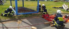 Strażacy podnoszą stalową konstrukcję kontenera przy użyciu poduszek pneumatycznych. Jeden z ochotników steruje poduszkami, natomiast trzech pozostałych zabezpiecza konstrukcje przez podkładanie drewnianych kantówek i klinów.