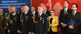 Na trybunie honorowej stoją zaproszeni goście mundurowi ubrani w mundury osoby cywilne w garnitury. W tle czerwony banner Państwowej Straży Pożarnej oraz granatowy baner Związku Ochotniczych Straży Pożarnych.