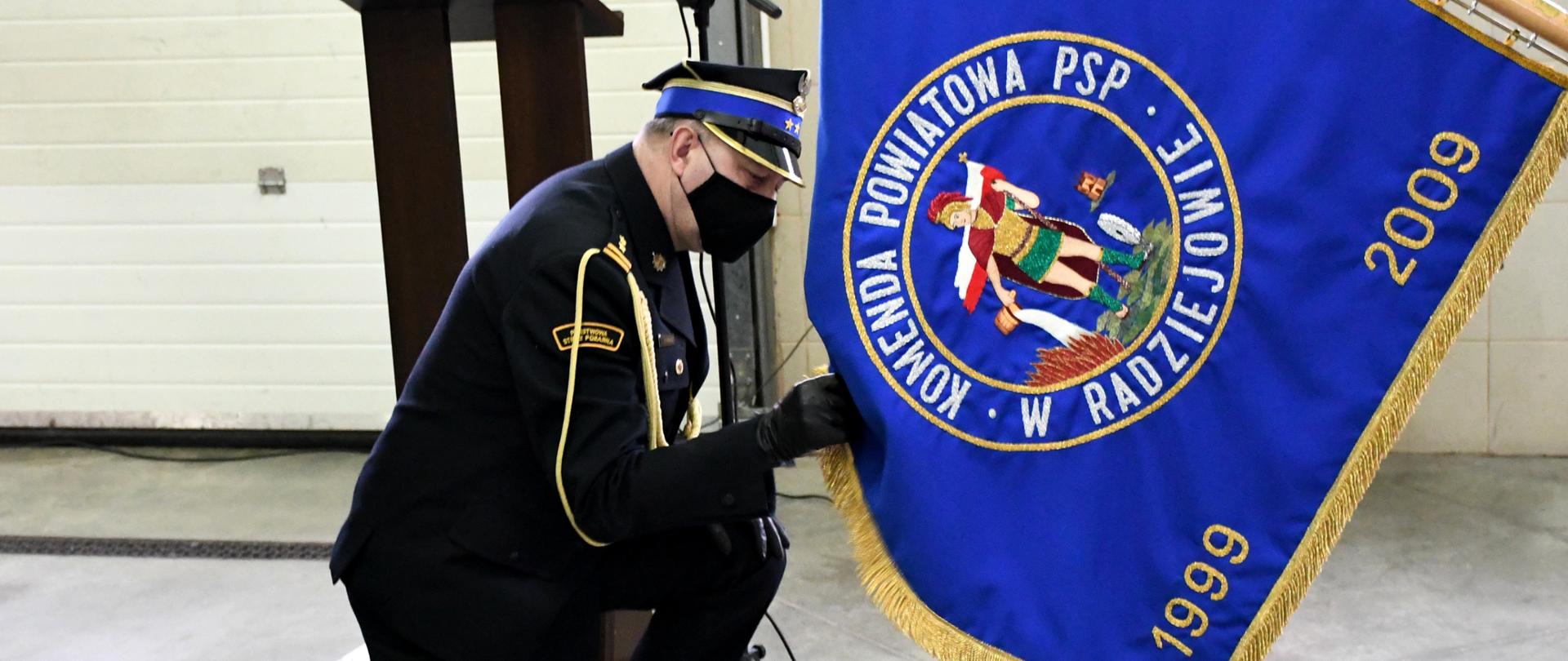 Zdjęcie przedstawia garaż Jednostki Ratowniczo-Gaśniczej Komendy Powiatowej Państwowej Straży Pożarnej w Radziejowie, oraz zastępcę komendanta powiatowego podczas żegnania się ze sztandarem Komendy Powiatowej PSP w Radziejowie