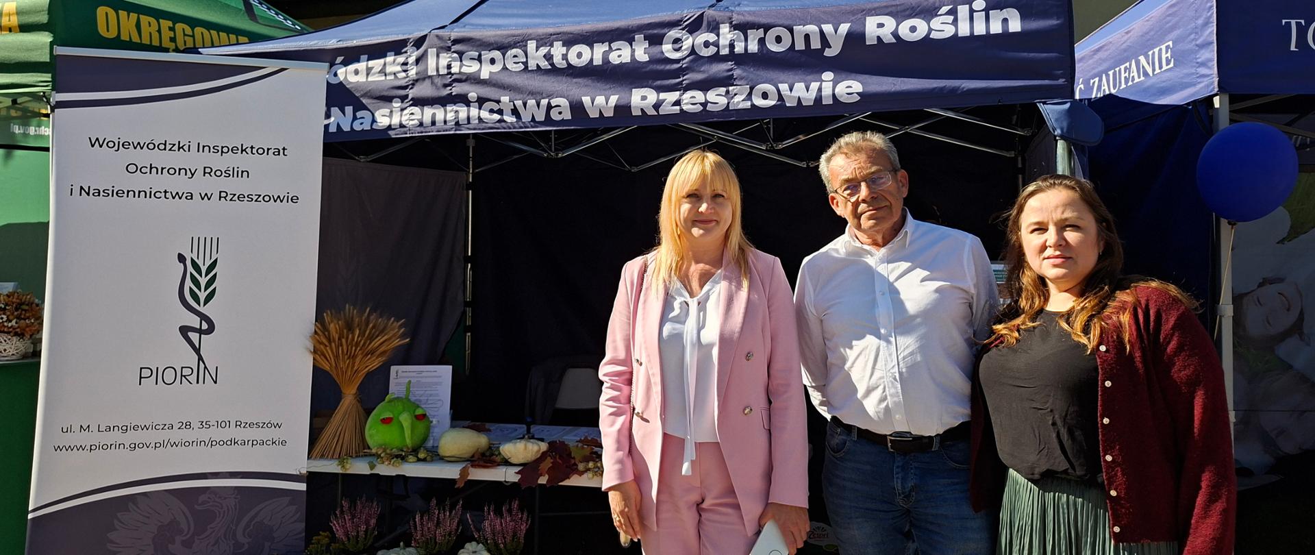 Trzy osoby stojące na chodniku. Stoją przed granatowym namiotem z napisem "Wojewódzki Inspektorat Ochrony Roslin i Nasiennictwa w Rzeszowie". Pod namiotem jest stolik a na nim różne materiały, ulotki. Stoisko ozdobione jest kwiatami i warzywami. Po lewej stronie stoi baner instytucji.