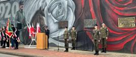 Teren przed Zakładem Karnym w Rawiczu. Na tle muru więziennego, na którym znajduje się mural, na chodniku stoją od lewej: poczty sztandarowe, strażak prowadzący uroczystość oraz wartownicy. Jest tam również stojak z flagami narodowymi oraz mównica. Na murze zawieszone sa trzy tablice pamiątkowe. 