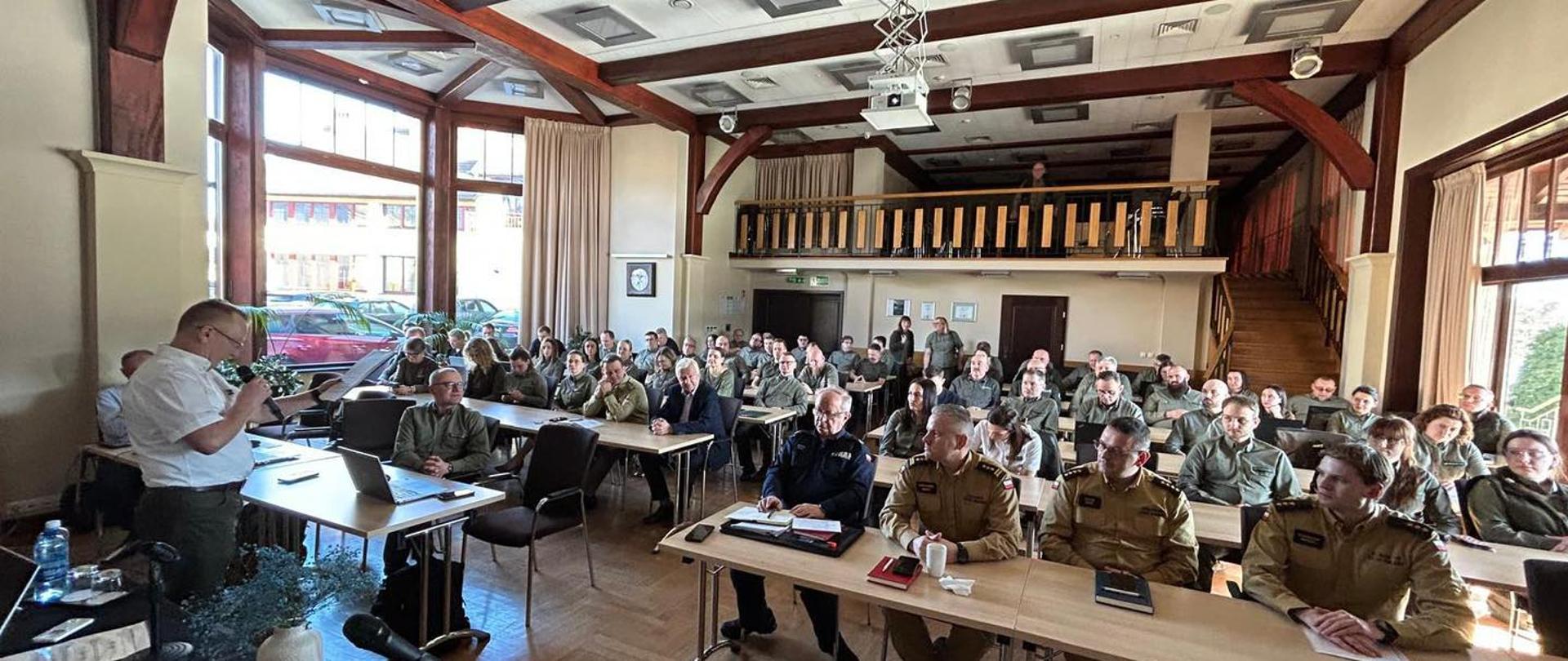 W dużej, jasnej sali konferencyjnej siedzi kilkadziesiąt osób, wiele z nich w mundurach. Uczestnicy siedzą przy długich stołach i słuchają mężczyzny stojącego z przodu przy mikrofonie i laptopie. Na suficie widać projektor, a z lewej strony duże okna wpuszczają światło dzienne.