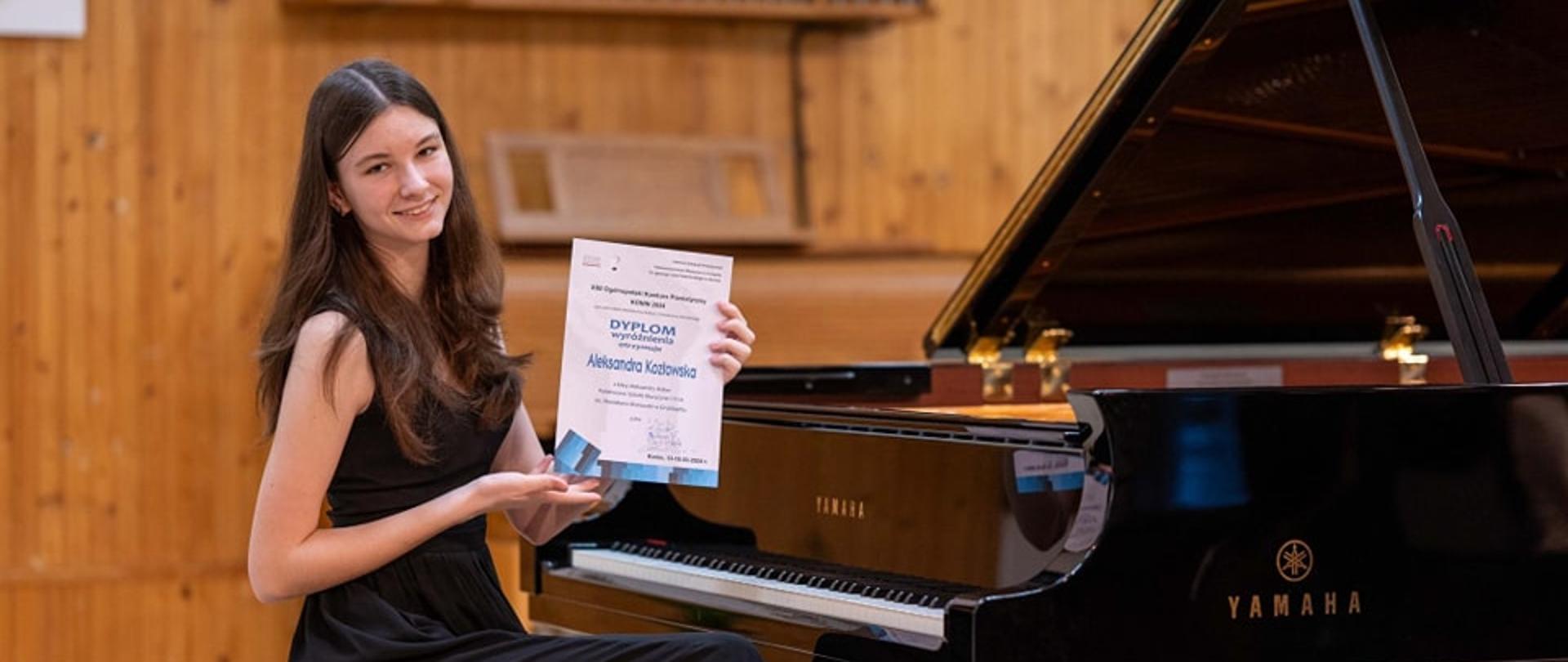 Uczennica na scenie przy fortepianie prezentuje zdobyty dyplom