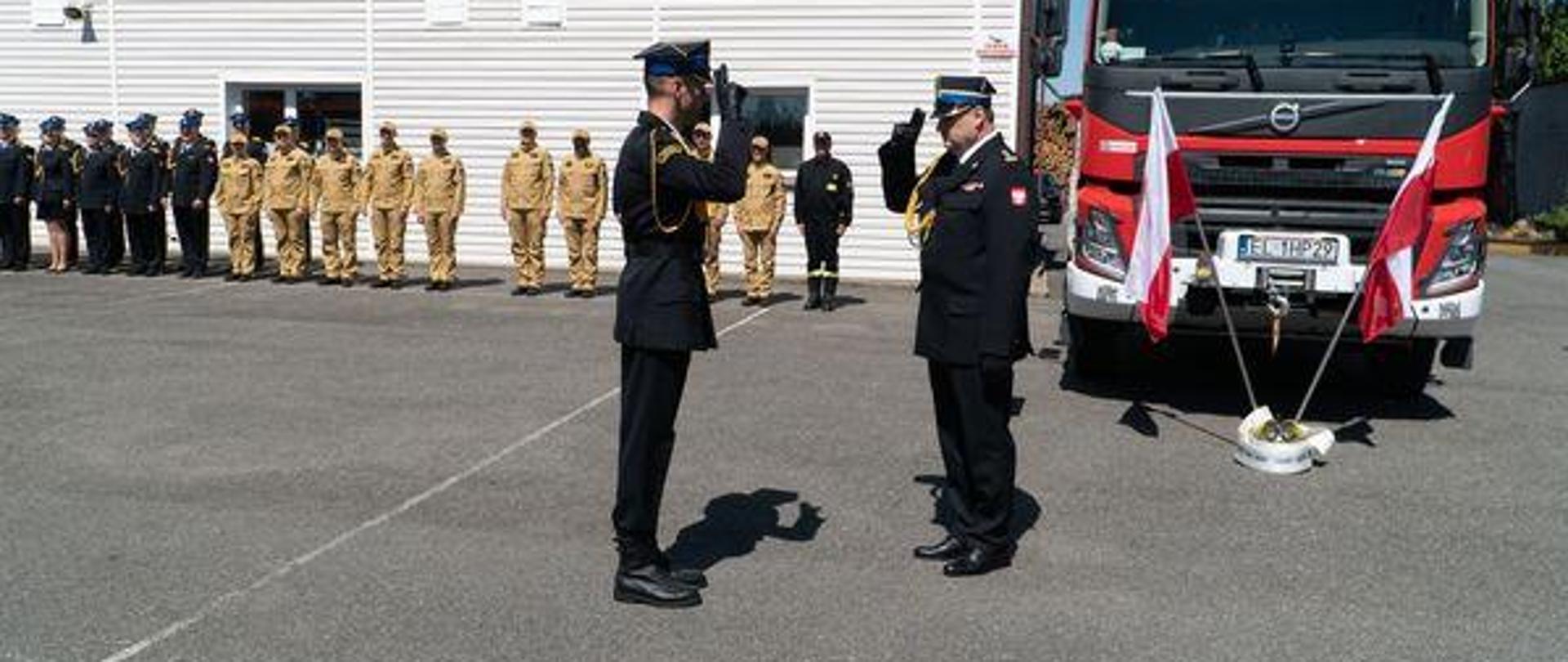 Na pierwszym planie dwóch strażaków salutujących do siebie z tyłu po prawej samochód gaśniczy z tyłu po lewej na tle budynku strażacy stojący na zbiórce