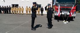 Na pierwszym planie dwóch strażaków salutujących do siebie z tyłu po prawej samochód gaśniczy z tyłu po lewej na tle budynku strażacy stojący na zbiórce