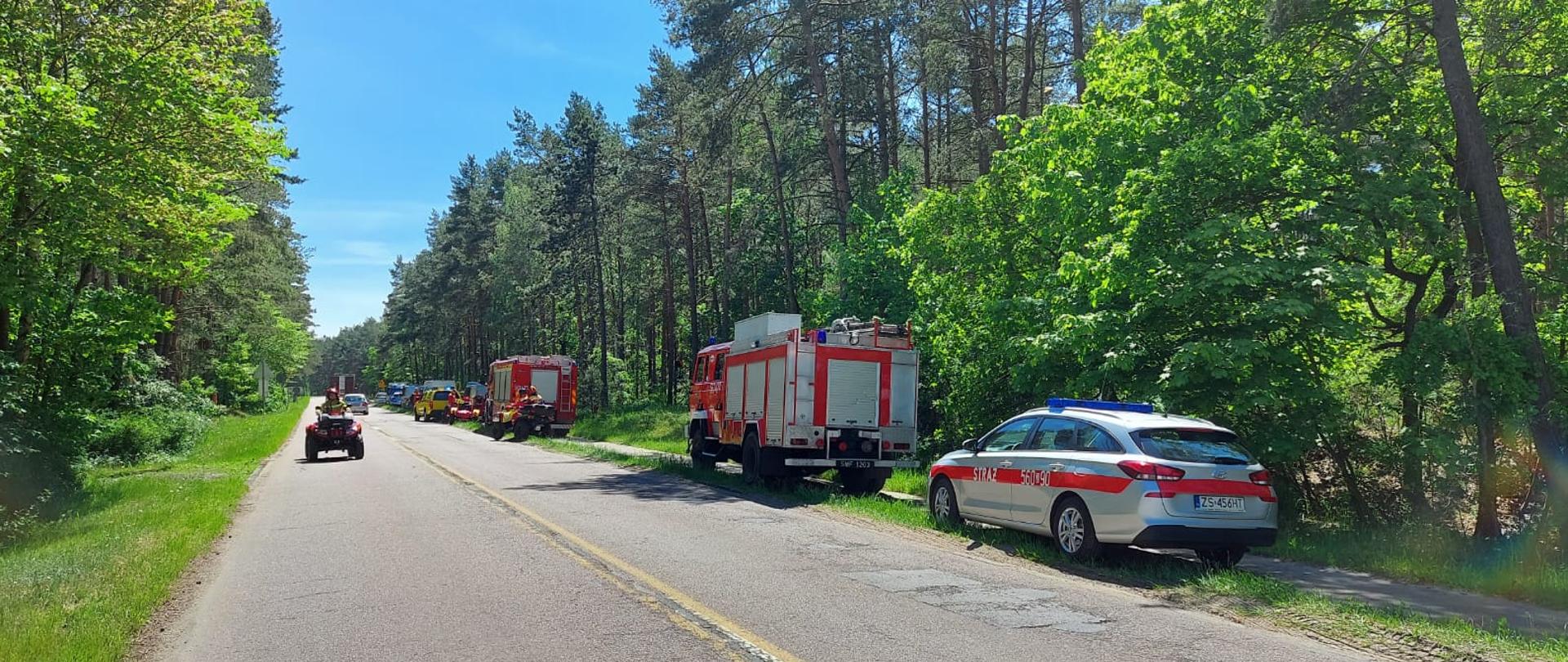 Ul. Karsiborska, po obu jej stronach las zielony. Po prawej stronie na poboczu stoją samochody służ. Na pierwszym planie samochód operacyjny, dalej 2 samochody gaśnicze. W oddali pojazdy WOPR, Policji i strażak na quadzie.