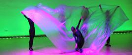 Wielokolorowe zdjęcie tancerek na scenie przed seledynowo - zielonym tłem.
