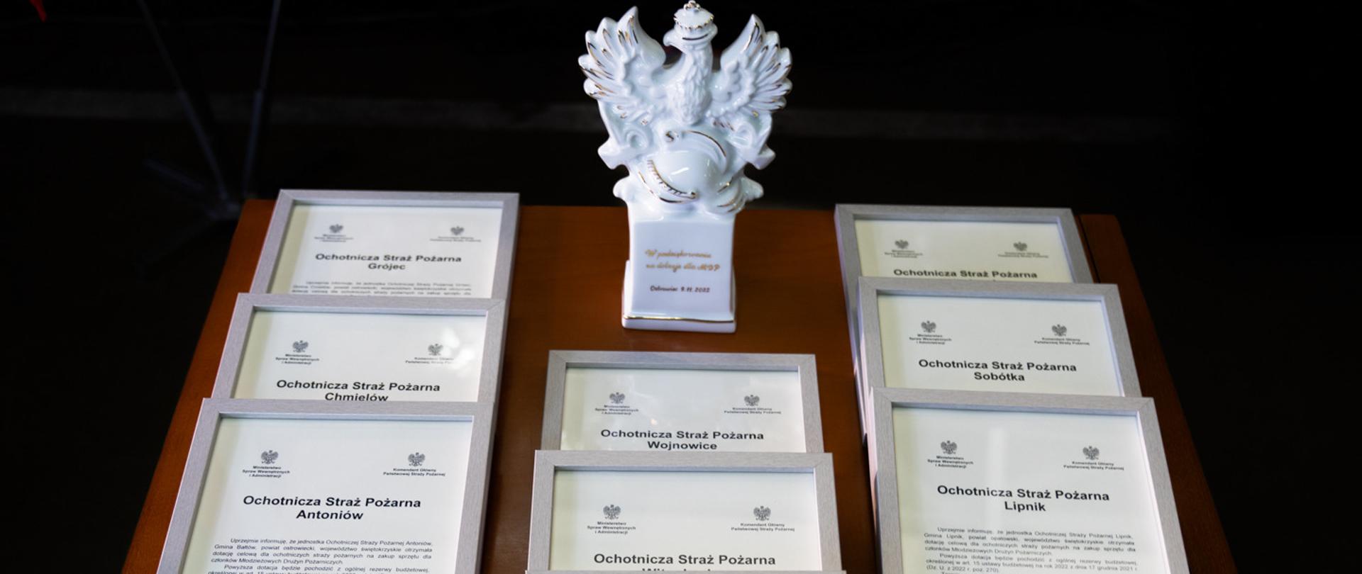 Na zdjęciu na drewnianym stoliku leży 9 decyzji o przyznaniu promes dla MDP. Decyzje te są oprawione w białe ramki. Na środku stolika stoi porcelanowy orzeł z grawerem dla komendanta głównego PSP.