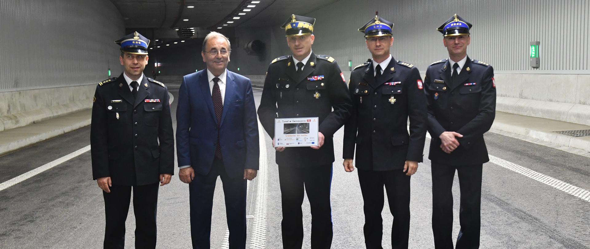 Zdjęcie przedstawia Prezydenta Miasta Świnoujście wraz z funkcjonariuszami PSP. od lewej: Zastępca Zachodniopomorskiego Komendanta PSP st. bryg. Mirosław Pender, Prezydent Miasta Janusz Żmurkiewicz, Zachodniopomorski Komendant PSP nadbryg. Jarosław Tomczyk, st. bryg. Robert Tarczyński, Komendant Miejski PSP w Świnoujściu st. kpt. Marek Wieczorek.
Zebrani stoją na jezdni tunelu. Funkcjonariusze w umundurowaniu wyjściowym. Granatowe mundury z rogatywkami.
