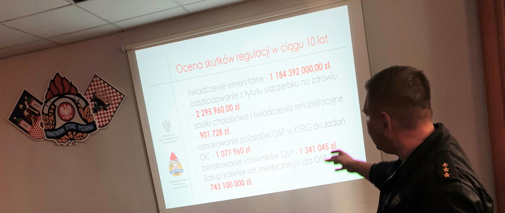 Na zdjęciu widać funkcjonariusza PSP ubranego w czarny mundur dowódczo-sztabowy. Człowiek ten prezentuje zarys ustawy, który jest wyświetlany za pomocą rzutnika na ekranie projekcyjnym. Obok ekranu projekcyjnego, na żółtej ścianie znajduje się logo PSP, herb miasta Kalisza i powiatu kaliskiego. Na suficie białe kasetony. Przed funkcjonariuszem komputer i brązowy stolik. Przed funkcjonariuszem stoi krzesło, na którym znajdują się dokumenty na kartkach A4.