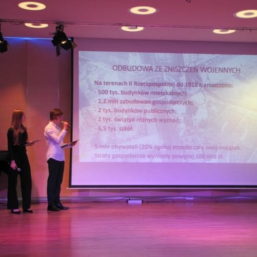 zdjęcie przedstawia uczniów szkoły prezentujących informacje o historii szkoły na ekranie multimedialnym na scenie sali koncertowej