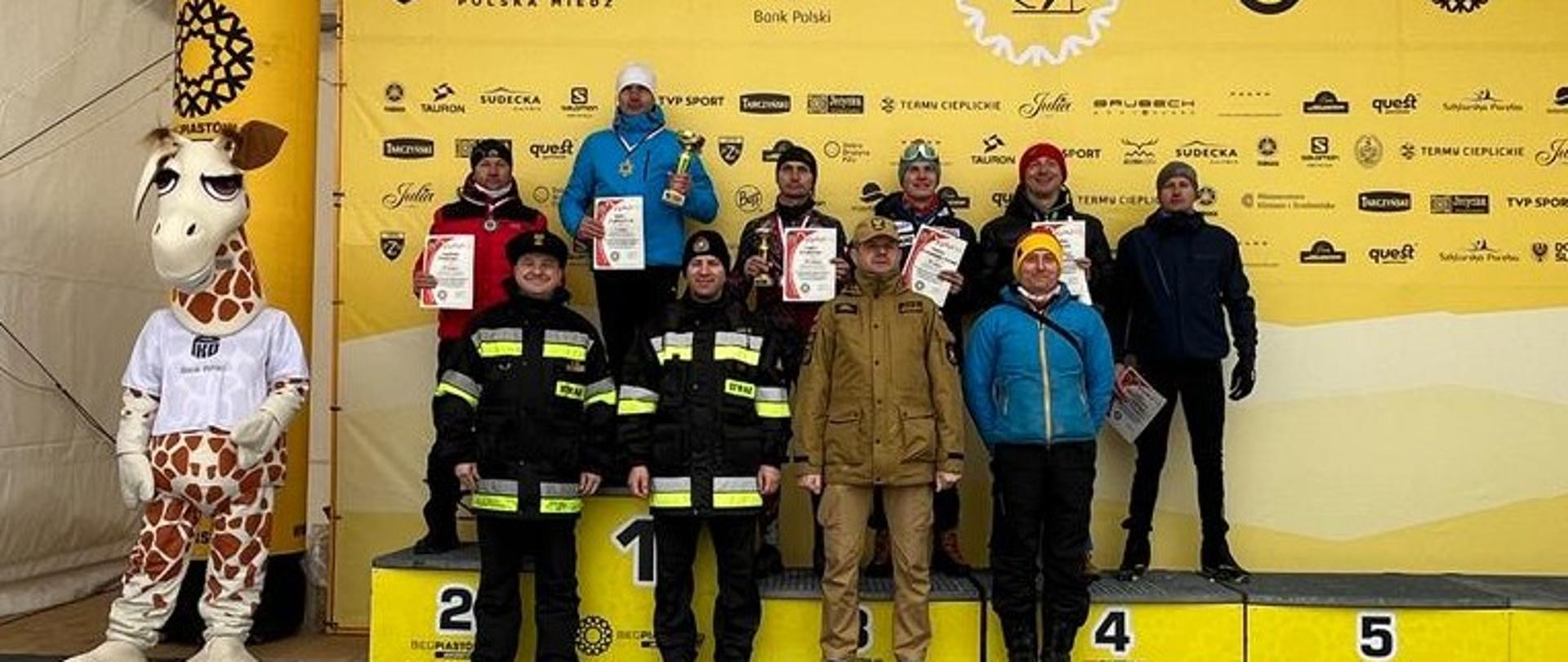 Na zdjęciu widać osoby stojące na scenie oraz podestach podium. Osoby ubrane są w umundurowanie strażackie oraz ubrania sportowe. Za osobami znajdującymi się na scenie znajduje się baner reklamowy koloru żółtego. Osoby znajdujące się na podium trzymają pucharu. Zdjęcie wykonane w dzień. 