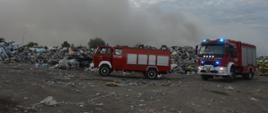 Pożar na składowisku odpadów. Plac składowy jednej z firm zajmującej się m.in. przetwarzaniem odpadów. Przed hałdą odpadów stoją dwa wozy strażackie z włączonymi sygnałami błyskowymi. Za drugim z aut widocznych jest kilku strażaków.