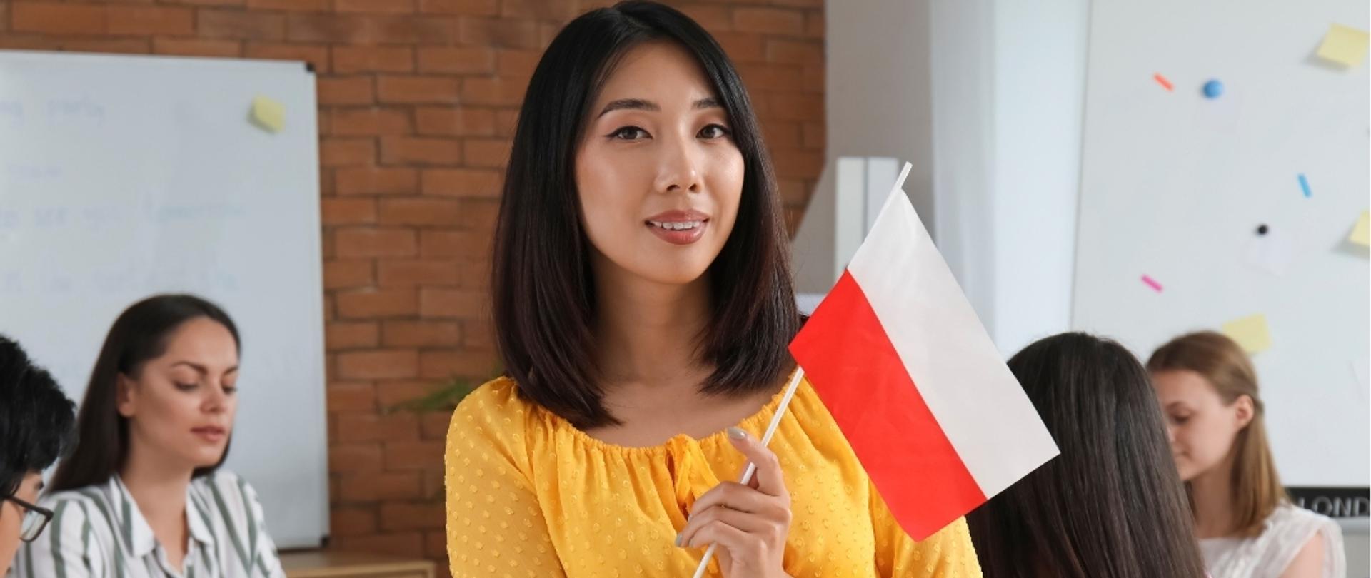 A woman standing in the middle is holding the Polish flag in her hand. In the background, several students can be seen sitting at a table.