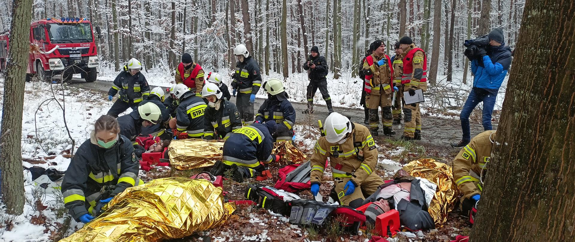 Symulowane działania ratownicze: poszkodowani wydobyci z pojazdów zgrupowani w jednym miejscu, na poboczu drogi. Strażacy OSP udzielają im pomocy. Poszkodowani na deskach ortopedycznych i owinięci folia izotermiczną. Strażacy OSP w ubraniach specjalnych i hełmach. Na drugim planie, na drodze strażacy PSP w czerwonych kamizelkach rozjemców obserwują i oceniają ćwiczących. Na drodze widać również nagrywającego wydarzenie kamerzystę TVP oraz fotografa PSP. W tle zimowa sceneria lasu i pojazd pożarniczy w lewym górnym rogu zdjęcia. 