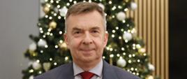 Minister Wieczorek stoi na tle udekorowanej bombkami i łańcuchami choinki.